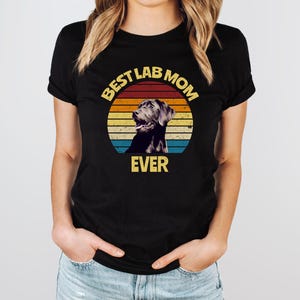 May include: A black short-sleeved t-shirt featuring a circular graphic with a retro sunset design in yellow, orange, and blue stripes. The graphic includes a black Labrador retriever illustration and the yellow text BEST LAB MOM EVER.