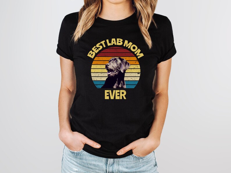 May include: A black short-sleeved t-shirt featuring a circular graphic with a retro sunset design in yellow, orange, and blue stripes. The graphic includes a black Labrador retriever illustration and the yellow text BEST LAB MOM EVER.