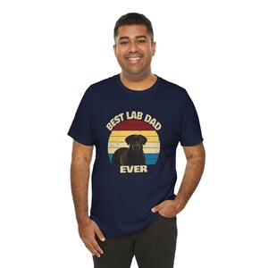 May include: Navy blue t-shirt with a circular graphic featuring a black Labrador Retriever and the text "Best Lab Dad Ever" in a retro style. The graphic has a sunset-like design with red, yellow, and blue stripes.