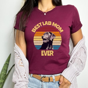 May include: A maroon short-sleeved t-shirt featuring a graphic of a black Labrador retriever inside a circular sunset design with horizontal stripes in yellow, orange, and blue. The text "BEST LAB MOM EVER" is printed in yellow block letters.