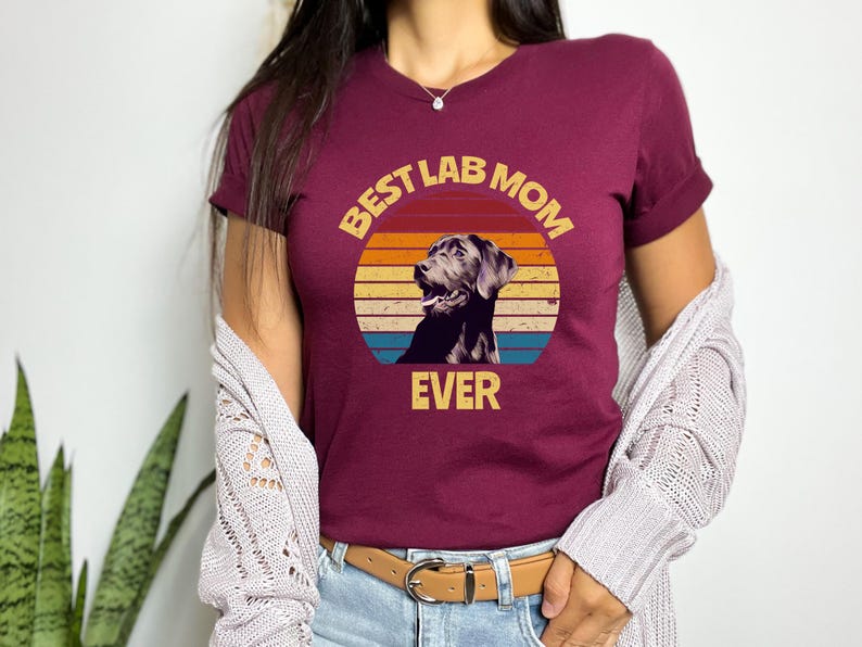 May include: A maroon short-sleeved t-shirt featuring a graphic of a black Labrador retriever inside a circular sunset design with horizontal stripes in yellow, orange, and blue. The text "BEST LAB MOM EVER" is printed in yellow block letters.
