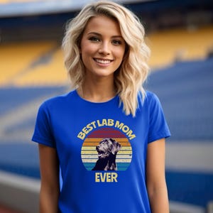 May include: A royal blue short-sleeve t-shirt featuring a circular graphic with a retro sunset design in orange, yellow, and blue stripes. The graphic includes a black Labrador retriever illustration and the text BEST LAB MOM EVER in yellow.