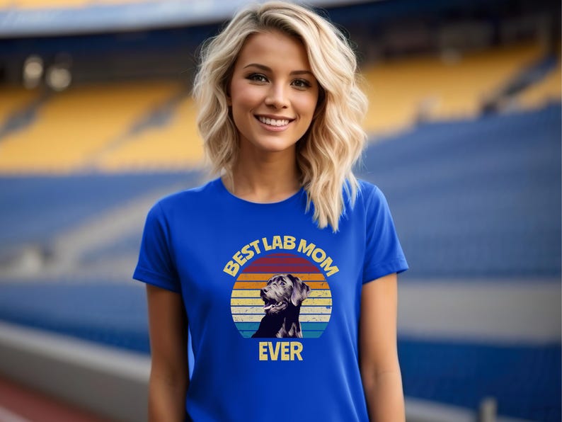 May include: A royal blue short-sleeve t-shirt featuring a circular graphic with a retro sunset design in orange, yellow, and blue stripes. The graphic includes a black Labrador retriever illustration and the text BEST LAB MOM EVER in yellow.