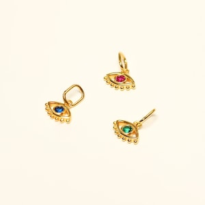 May include: Three gold evil eye charms with different colored gemstones. The charms are in the shape of an eye with a circle in the center and a fringe of small beads around the edge. The gemstones are blue, green, and pink.