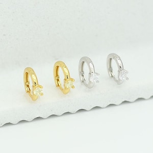 May include: Three pairs of small hoop earrings with a single clear stone set in each hoop. The hoops are available in gold and silver.