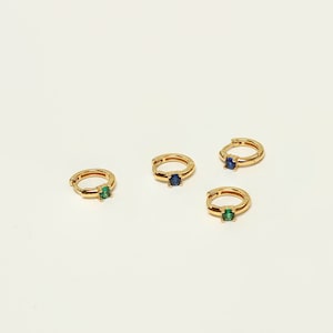Pode incluir: Quatro brincos de argola de ouro com pequenas pedras preciosas. Dois brincos têm uma pedra preciosa azul e dois brincos têm uma pedra preciosa verde.
