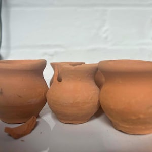 May include: A collection of small, unglazed terracotta pots, each with a rounded shape and a wide rim. One pot has a broken fragment nearby. The pots are a warm orange-brown colour, set against a white brick wall.