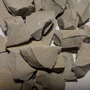 May include: A collection of light grey clay pieces with irregular shapes and rough, fractured surfaces. The chunks vary in size and texture, with some showing a slightly lighter colour. The clay appears dry and earthy.