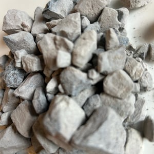 May include: A collection of light grey, irregularly shaped rocks with a slightly rough texture. Some rocks have hints of blue-grey. The rocks are clustered together, creating a natural, earthy appearance.
