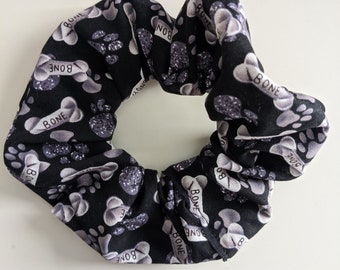 May include: Black and white scrunchie with a paw print and bone pattern. The scrunchie has the word "BONE" repeated throughout the pattern.