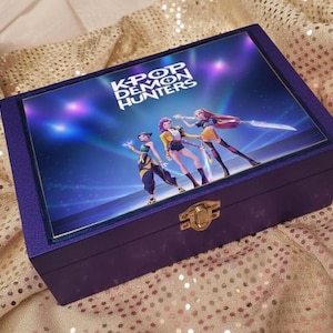 May include: A purple wooden box with a gold clasp. The lid features a colorful graphic of three anime-style characters with the text "K-POP DEMON HUNTERS" in white. The box is on a gold sequined surface.