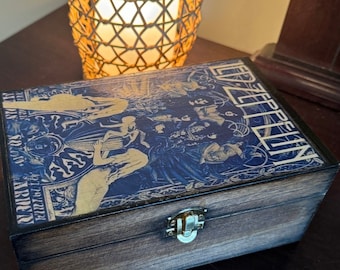 May include: A wooden box with a hinged lid and a metal latch. The box is decorated with a vintage-style image of a band, with the word "Led Zeppelin" printed on the side. The box is sitting on a dark wooden surface.