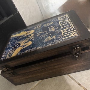 May include: A wooden box with a hinged lid, featuring a vintage design. The lid displays a blue and gold graphic with the text "ZEPPELIN". The box is stained a dark brown and has metal clasps.