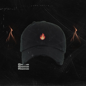 Może przedstawiać: Czarna czapka bejsbolówka z małym, pomarańczowym płomieniem wyszytym z przodu. Czapka ma przetarty wygląd i znajduje się na czarnym tle.