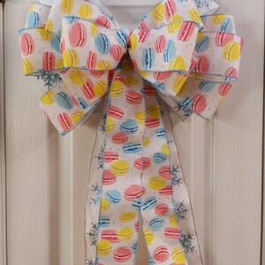 May include: A white bow with a macaron pattern in pink, yellow, and blue. The bow has a silver snowflake trim.