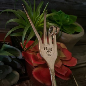 May include: A silver fork with the word "PEACE" and a peace sign engraved on the handle. The fork is bent into a peace sign shape.