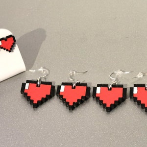 May include: Four pairs of pixel heart earrings. The earrings are red with black outlines and are shaped like pixelated hearts. The earrings are on a white surface.