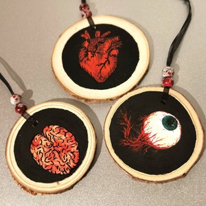 May include: Three round wooden ornaments with black backgrounds and red and white painted anatomical illustrations. The ornaments feature a heart, an eye, and a brain.
