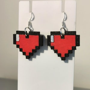 May include: A pair of red pixel heart earrings with black outlines and silver hooks. The hearts are made of wood and have a glittery finish.
