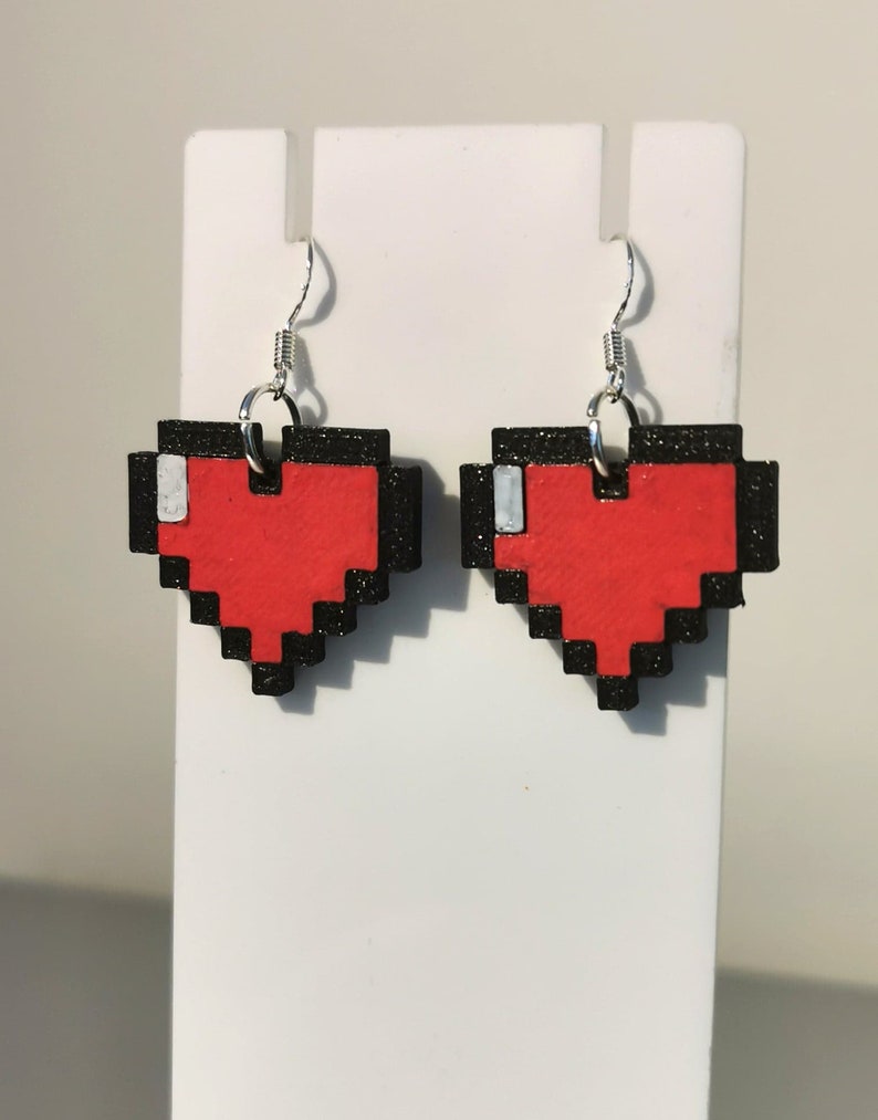 May include: A pair of red pixel heart earrings with black outlines and silver hooks. The hearts are made of wood and have a glittery finish.