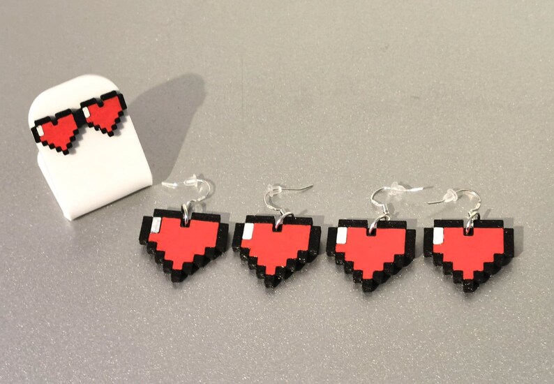 May include: Four pairs of pixel heart earrings. The earrings are red with black outlines and are shaped like pixelated hearts. The earrings are on a white surface.