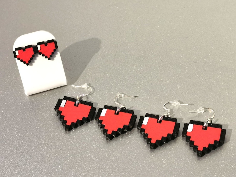 May include: Four pairs of pixel heart earrings. The earrings are red with black outlines and are shaped like hearts. The earrings are on a white display stand.