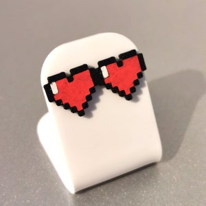 May include: A pair of red pixel heart earrings with black outlines. The earrings are displayed on a white stand.