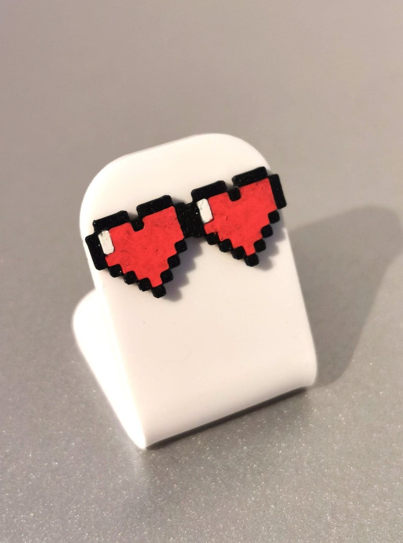 May include: A pair of red pixel heart earrings with black outlines. The earrings are displayed on a white stand.