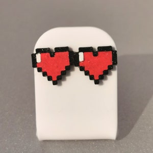 May include: A pair of red pixel heart earrings with black outlines. The earrings are on a white display stand.