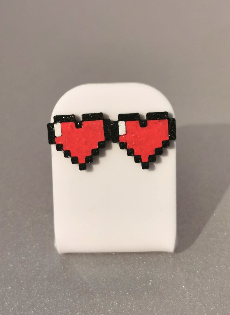 May include: A pair of red pixel heart earrings with black outlines. The earrings are on a white display stand.