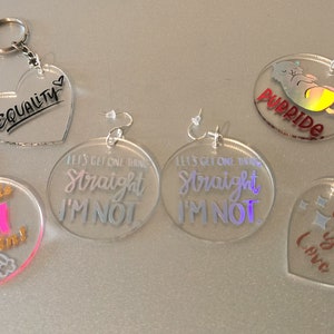May include: Assortment of acrylic keychains and earrings with LGBTQ+ themed designs. Keychains include a heart with "EQUALITY" written on it, a circle with "Sounds Gay I'm in!" and a rainbow, and a circle with a cat and the word "Purride". Earrings are a pair of circles with "Let's get one thing straight I'm not" written on them.