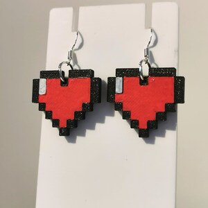 May include: A pair of pixelated heart earrings with black outlines and red centers. The earrings are hanging from silver hooks.