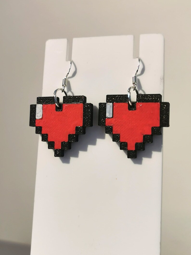 May include: A pair of pixelated heart earrings with black outlines and red centers. The earrings are hanging from silver hooks.