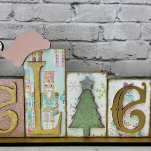 May include: A wooden sign that spells out the word "BELIEVE" in gold letters. The letters are on individual blocks with various patterns and a Santa hat on top. The sign is on a wooden base.