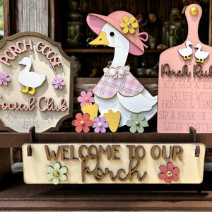 May include: Wooden porch decor featuring a goose theme. Includes a sign that says "Porch Goose Social Club", a goose figurine, and a sign that says "Welcome to Our Porch". The decor is adorned with flowers and the sign "Porch Rules".