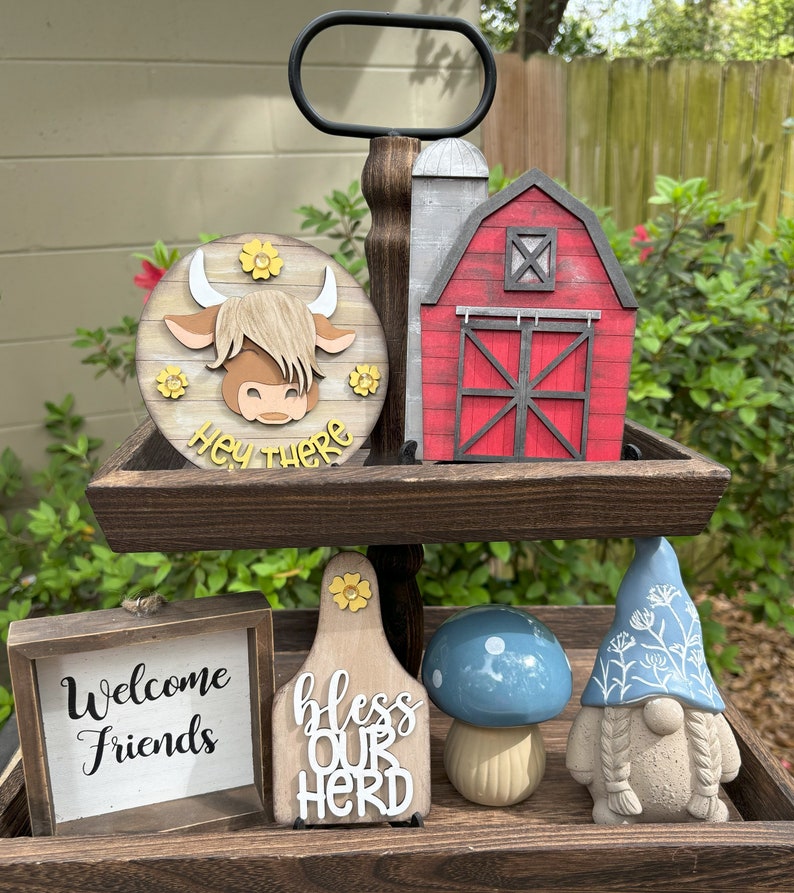 Puede incluir: Una bandeja de madera de dos niveles con decoraciones con tem&aacute;tica de granja. Incluye un letrero redondo con la cara de una vaca y el texto "Hey There", un granero rojo, un letrero que dice "Welcome Friends", una etiqueta de vaca que dice "Bless Our Herd", un hongo azul y un gnomo.