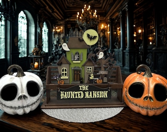 May include: Halloween-themed decor featuring a haunted house centerpiece with ghosts, bats, and pumpkins. The centerpiece has the text "The Haunted Mansion". Flanking the house are two carved pumpkin heads, one white and one orange.