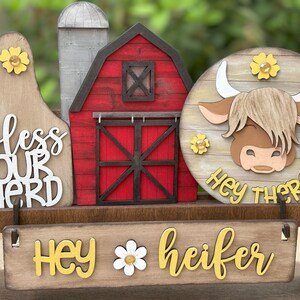 Puede incluir: Decoraci&oacute;n de madera con tem&aacute;tica de granja, con un granero rojo, un silo y un letrero redondo con una vaca de las Highlands. Los letreros dicen "Hey there" y "Hey heifer" con acentos de margaritas. El letrero de la izquierda dice "bless our herd".