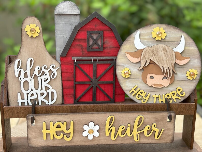 Puede incluir: Decoraci&oacute;n de madera con tem&aacute;tica de granja, con un granero rojo, un silo y un letrero redondo con una vaca de las Highlands. Los letreros dicen "Hey there" y "Hey heifer" con acentos de margaritas. El letrero de la izquierda dice "bless our herd".