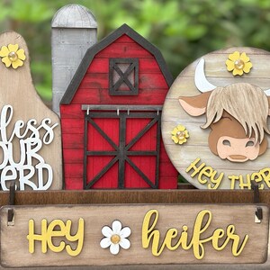 Puede incluir: Decoraci&oacute;n de madera con tem&aacute;tica de granja con un granero rojo, un silo y un letrero redondo con un dise&ntilde;o de vaca de las Tierras Altas. El letrero dice "Hey there" y "Hey heifer". La decoraci&oacute;n incluye el texto "bless our herd".