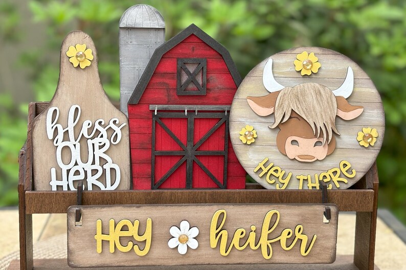 Puede incluir: Decoraci&oacute;n de madera con tem&aacute;tica de granja con un granero rojo, un silo y un letrero redondo con un dise&ntilde;o de vaca de las Tierras Altas. El letrero dice "Hey there" y "Hey heifer". La decoraci&oacute;n incluye el texto "bless our herd".
