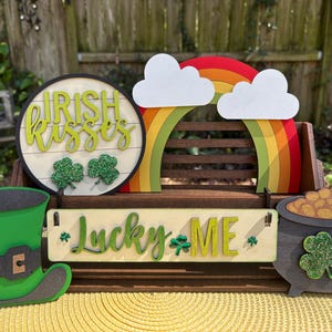 May include: A wooden sign with the text "Irish Kisses" in green letters, a rainbow with clouds, a pot of gold, a green leprechaun hat, and a sign that says "Lucky Me" with shamrocks.