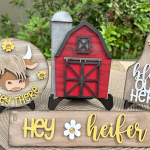 Puede incluir: Decoraciones de madera con tem&aacute;tica de granja. Incluye un letrero redondo con la cara de una vaca y el texto "Hey There", un granero rojo, un letrero que dice "Hey Heifer" y un letrero que dice "Bless Our Herd".