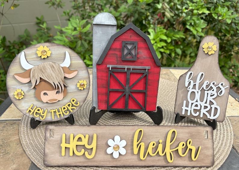 Puede incluir: Decoraciones de madera con tem&aacute;tica de granja. Incluye un letrero redondo con la cara de una vaca y el texto "Hey There", un granero rojo, un letrero que dice "Hey Heifer" y un letrero que dice "Bless Our Herd".