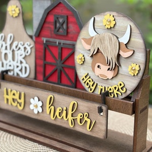 Puede incluir: Decoraci&oacute;n de madera con tem&aacute;tica de granja con una vaca, un granero y una campana de vaca. La vaca tiene una cara marr&oacute;n y cuernos blancos. El letrero dice "Hey there heifer" en letras amarillas. Se incluyen acentos de flores amarillas.