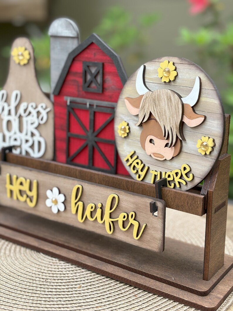 Puede incluir: Decoraci&oacute;n de madera con tem&aacute;tica de granja con una vaca, un granero y una campana de vaca. La vaca tiene una cara marr&oacute;n y cuernos blancos. El letrero dice "Hey there heifer" en letras amarillas. Se incluyen acentos de flores amarillas.