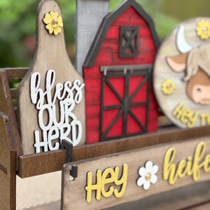 Puede incluir: Decoraci&oacute;n de madera con tem&aacute;tica de granja con un granero rojo, una etiqueta de vaca con "bless our herd" y un letrero redondo con una cara de vaca. El letrero tambi&eacute;n tiene el texto "Hey heifer" en amarillo y blanco. La decoraci&oacute;n incluye acentos de flores amarillas.
