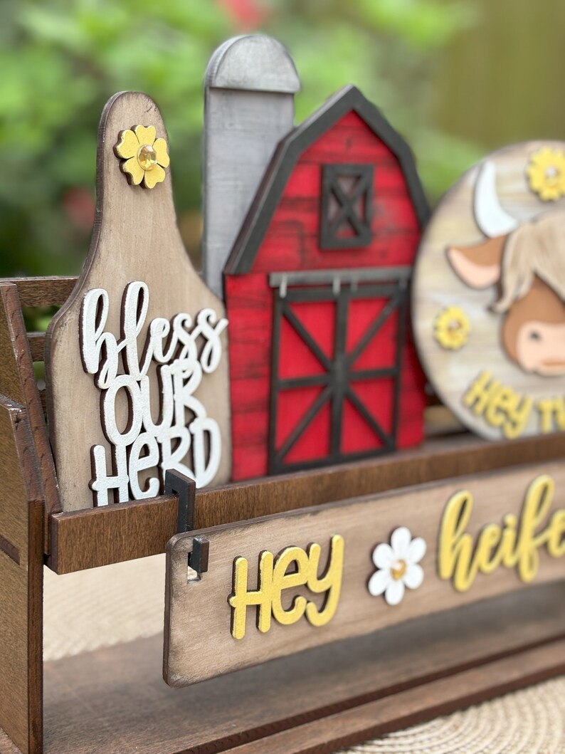 Puede incluir: Decoraci&oacute;n de madera con tem&aacute;tica de granja con un granero rojo, una etiqueta de vaca con "bless our herd" y un letrero redondo con una cara de vaca. El letrero tambi&eacute;n tiene el texto "Hey heifer" en amarillo y blanco. La decoraci&oacute;n incluye acentos de flores amarillas.