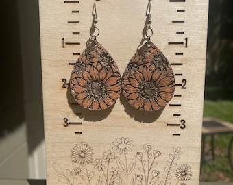 Könnte beinhalten: Holz-Tropfenohrringe mit Sonnenblumenmuster. Die Ohrringe sind hellbraun mit einem dunkleren braunen Sonnenblumenmuster. Auf dem Holzständer ist der Text "Autumn Gal Creations" zu sehen.