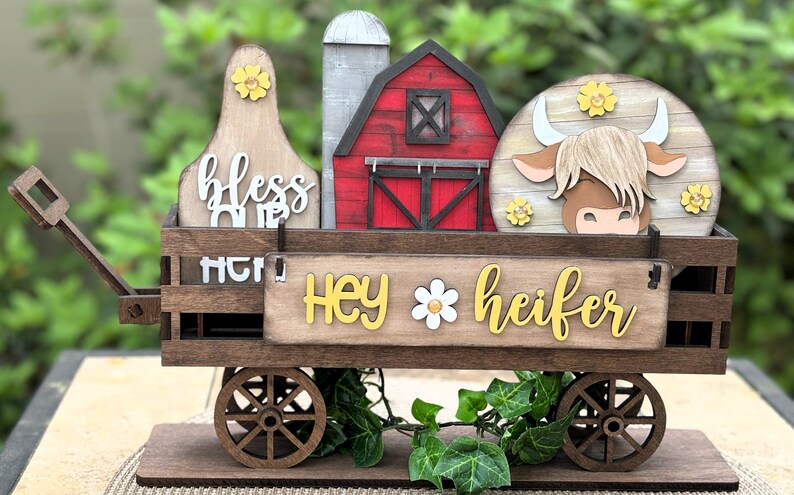 Puede incluir: Carro decorativo de madera con tem&aacute;tica de granja con un granero rojo, una vaca y un silo. El carro tiene las palabras "Hey heifer" y "bless our home". El carro es marr&oacute;n con detalles en amarillo y blanco y tiene ruedas de madera.